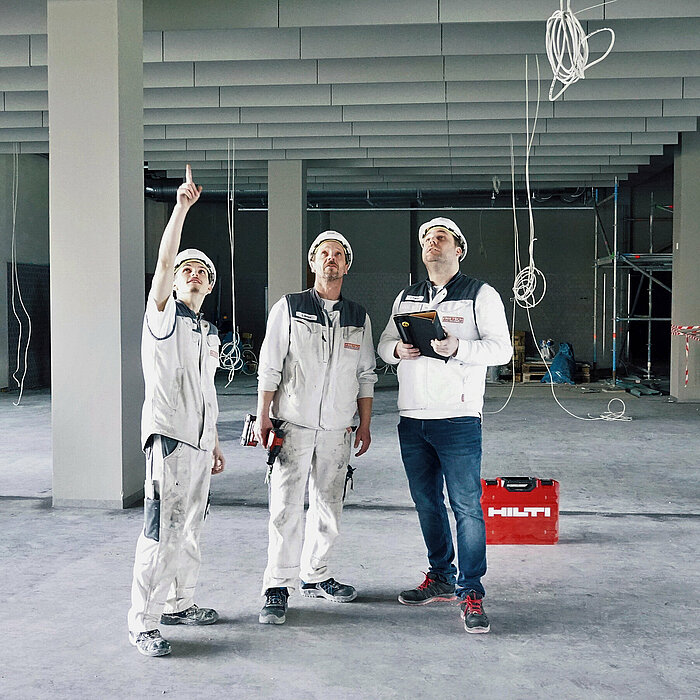 Photo: Hilti: 3 men dressed in white are inside a construction site looking at the ceiling, a Hilti tool case is in the back