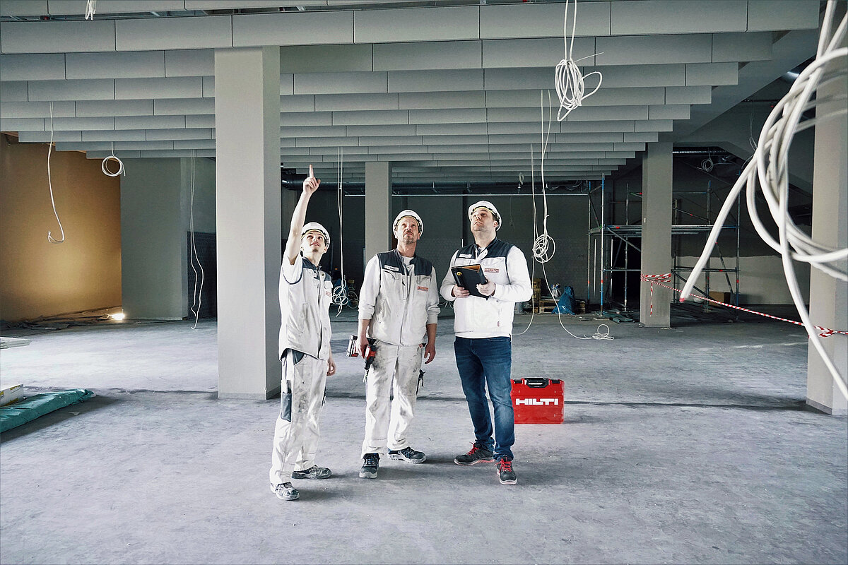Photo: Hilti: 3 men dressed in white are inside a construction site looking at the ceiling, a Hilti tool case is in the back
