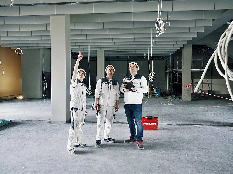 Photo: Hilti: 3 men dressed in white are inside a construction site looking at the ceiling, a Hilti tool case is in the back