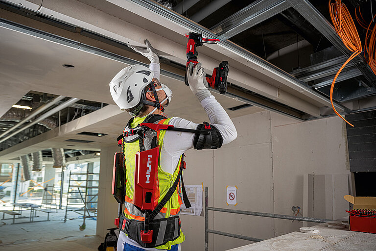 Photo: Hilti: A man with a white helmet and a mask wearing the Hilti Exo-Skeleton and working overhead with a Hilti tool