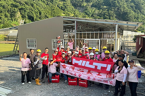 Photo: Hilti: Hilti Taiwan workers posing with the Hilti tool cases and the "Hilti" and "New Taiwan Volunteer Group" logo flags, an unfinished house behind them 