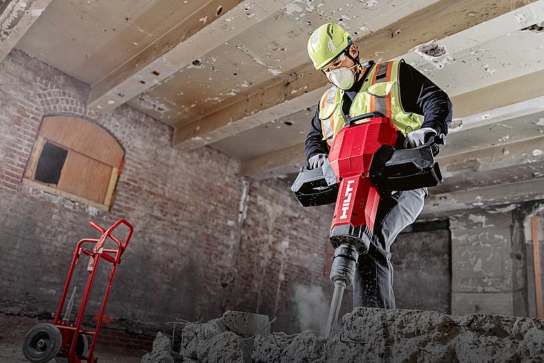 Hilti worker wearing a hard hat, safety glasses, and high-visibility vest while working with a Hilti tool on a construction site.