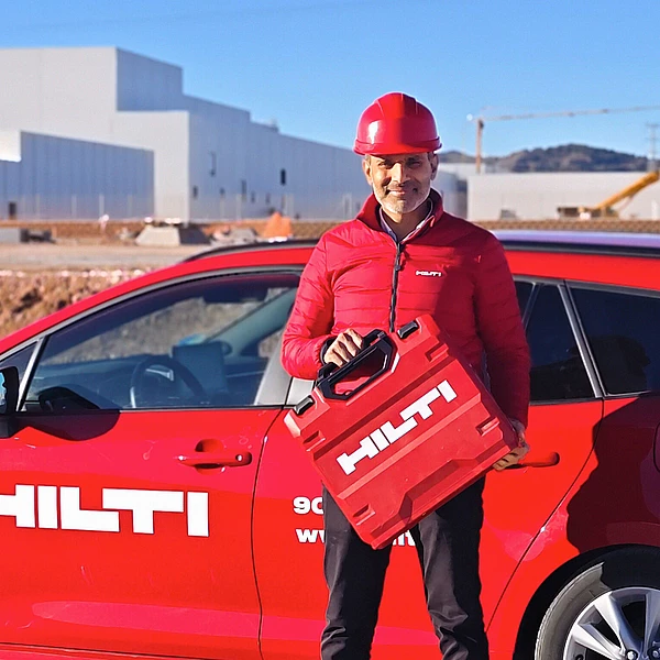 Akash Chauhan standing in front of a red Hilti car while wearing a safety helmet and a red Hilti jacket and holding a red toolbox.