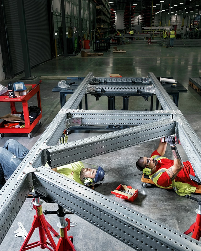 Two workers in a warehouse, wearing safety clothing and high-visibility vests, lying on the floor and screwing into a modular support system from below.