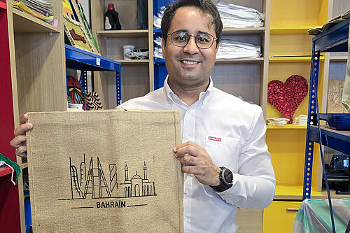 A Hilti volunteer wearing a white shirt smiles at the camera while holding a jute bag with the word “Bahrain” printed on it.
