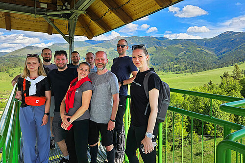 Group photo of hikers standing under a wooden shelter, with green mountains in the background.