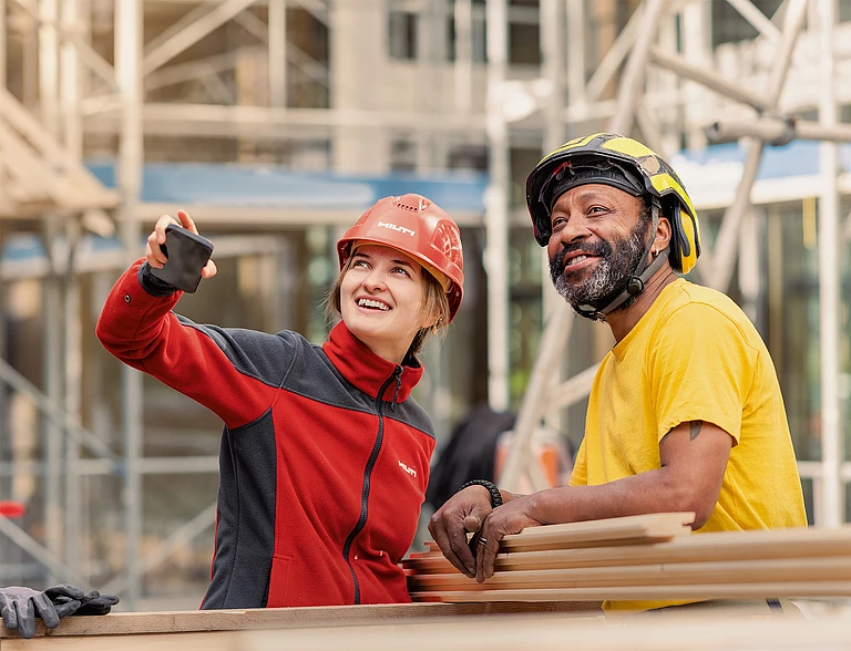 A Hilti salesperson in Hilti workwear and a safety helmet points ahead while standing next to a customer in a yellow T-shirt and a safety helmet. They appear to be discussing or inspecting something on a construction site.