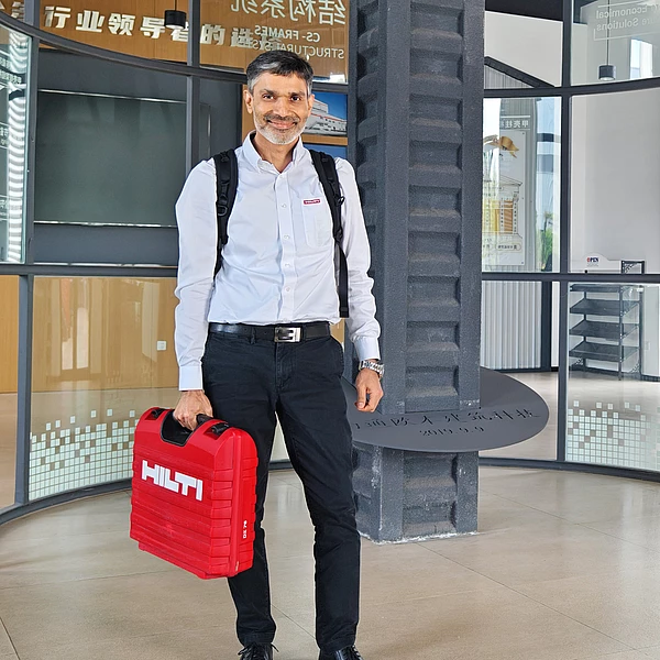 Akash Chauhan in a white shirt and black trousers, wearing a backpack while holding a red Hilti-toolbox.