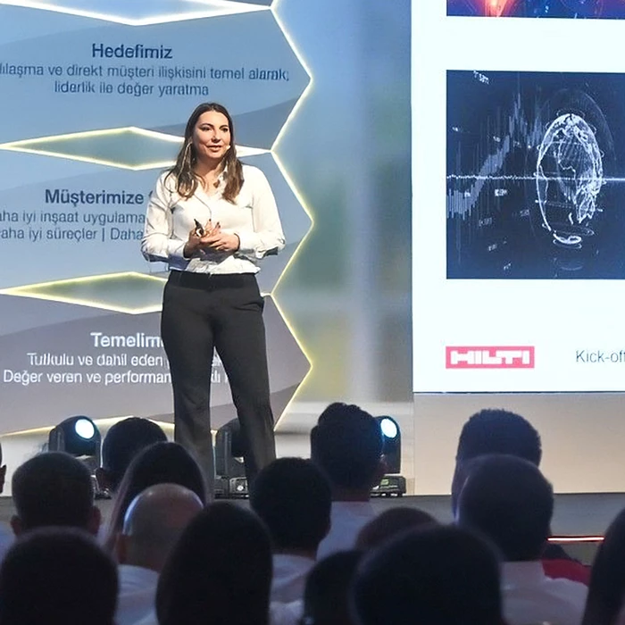 Banu Deniz Çetinkol gives a speech to colleagues on a stage while wearing black pants and a white shirt.