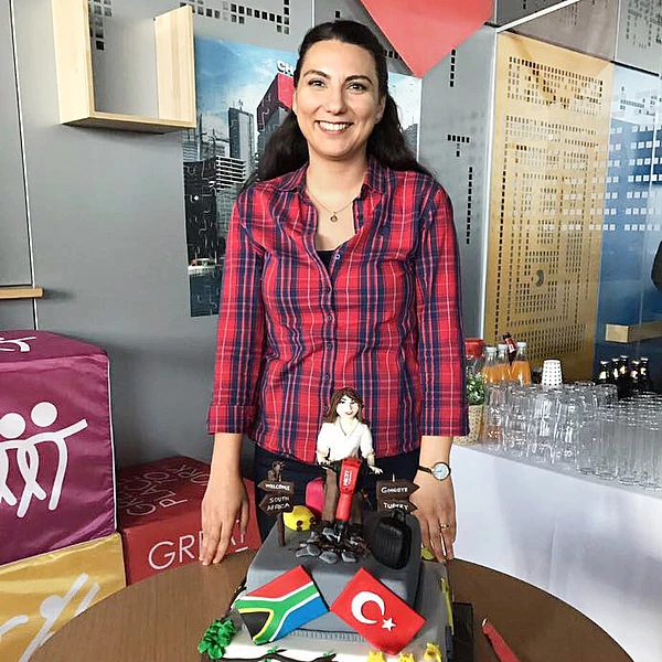 Banu Deniz Çetinkol smiling and wearing a red checkered shirt. In front of her is a themed cake depicting her as a marzipan figure, with the flags of Turkey and South Africa underneath. 